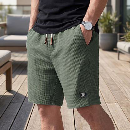Mann in grüner Waffelstruktur-Short mit hellem Kordelzug, Armbanduhr und Hand in der Tasche, auf einer sonnigen Holzterrasse. 5