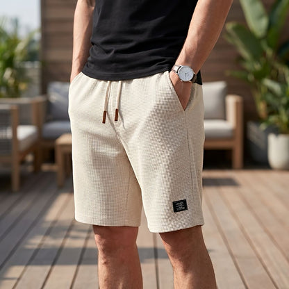 Mann in beiger Short mit Waffelstruktur, Kordelzug und Lederenden, Hand in der Tasche, auf einer Holzterrasse mit Sitzmöbeln. 8