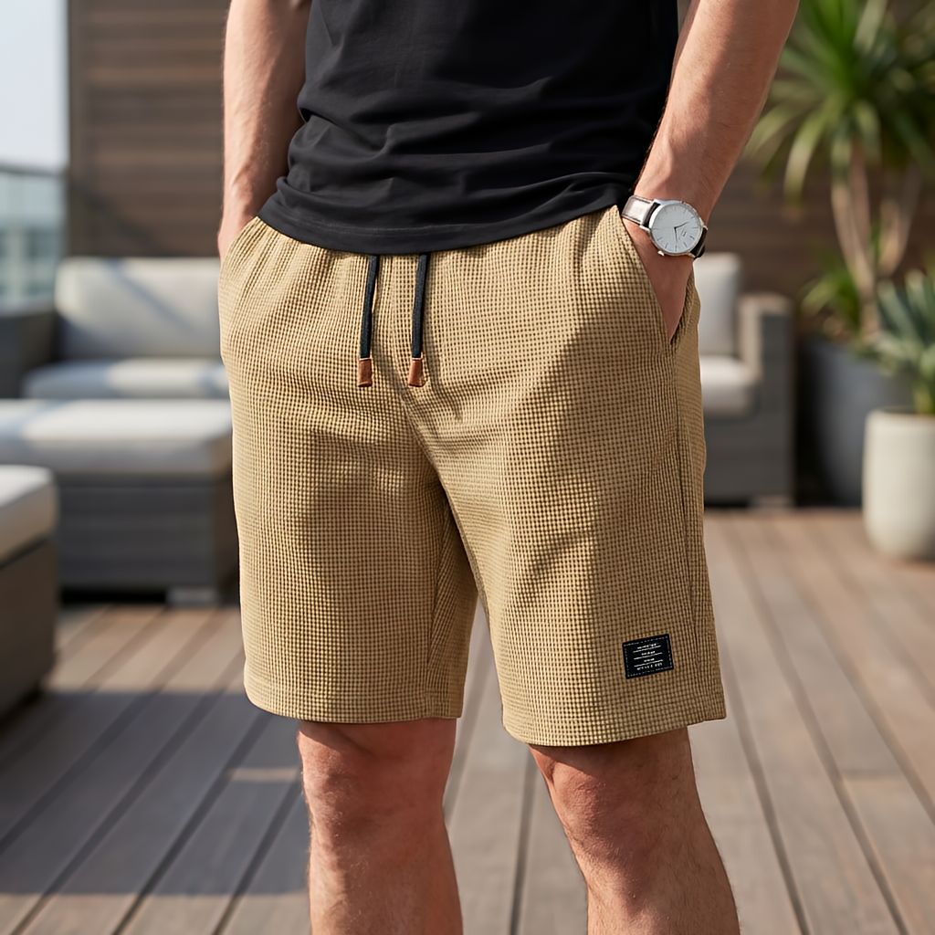 Mann in sandfarbener Waffelstruktur-Short mit dunklem Kordelzug und Lederenden, Hand in der Tasche, vor Loungemöbeln auf einer Dachterrasse. 4