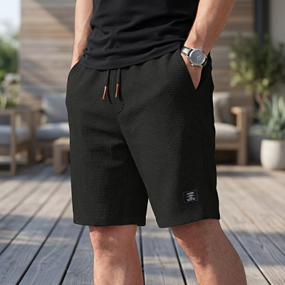 Mann in schwarzer Short mit Waffelstruktur, Kordelzug und kleinem Stoffetikett am Bein, Hand in der Tasche, auf einer Holzterrasse. 3