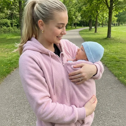 Frau mit Pferdeschwanz im rosa Fleece-Hoodie trägt ihr Baby mit blauer Mütze im Einsatz, auf einem Parkweg. 4