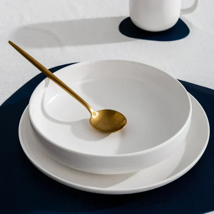 Weiße Keramikschale mit goldfarbenem Löffel auf blauem Untersetzer, modernes Geschirr-Set.