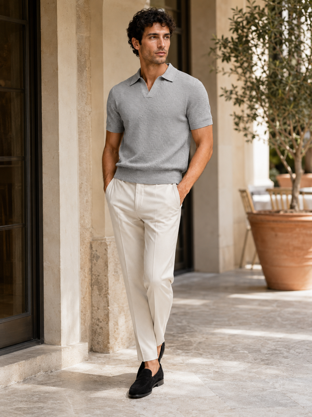 Model im grauen Strickpolo steht auf einer sonnigen Terrasse mit Olivenbaum, cremeweiße Hose, schwarze Loafer 30