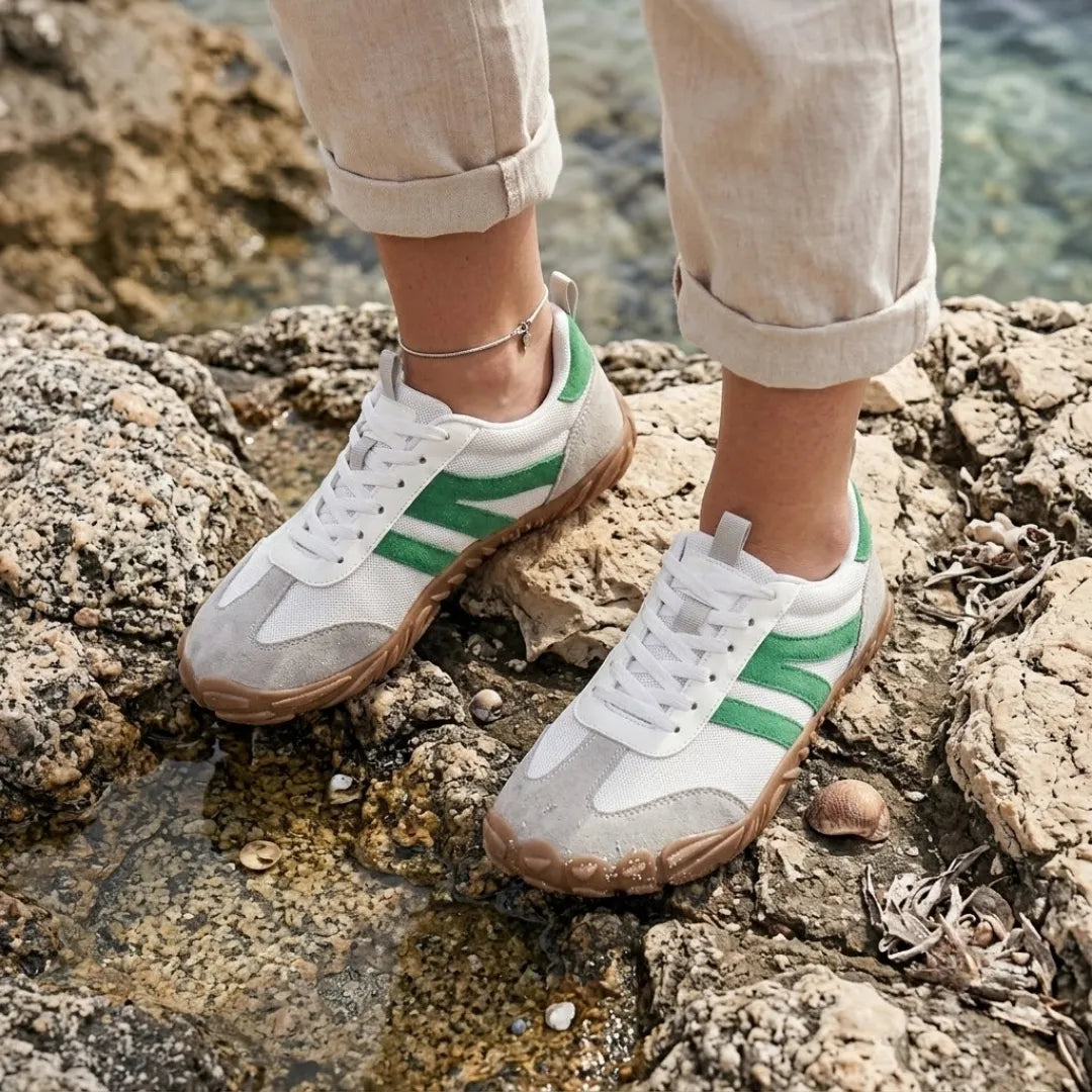 Frau in hellen Leinenhosen trägt weiß-grüne Barfußschuhe auf Felsen am Wasser, mit Fußkettchen und Muscheln im Hintergrund. 29