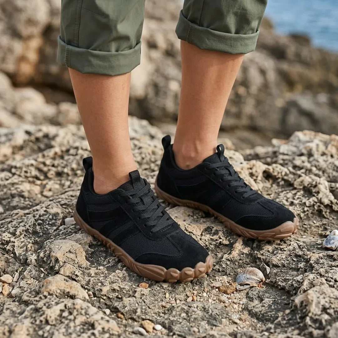 Schwarze Barfußschuhe, getragen mit hochgekrempelten grünen Hosen auf felsigem Küstengrund mit Muscheln und Meer im Hintergrund. 20