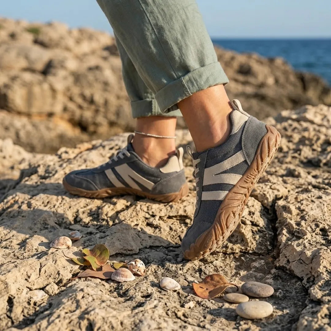 Graue Barfußschuhe mit beigen Streifen, getragen an Füßen mit Knöchelkettchen auf felsiger Küste mit Muscheln und Kieseln. 2