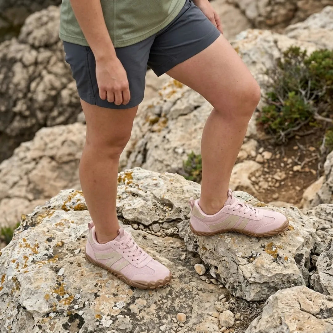 Rosafarbene Barfußschuhe mit beigen Streifen, getragen mit grauen Shorts auf Felsen mit Flechten und Büschen. 11