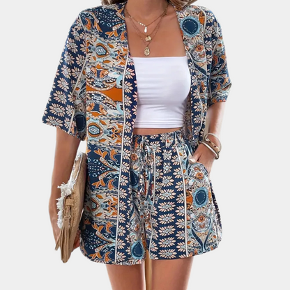 Frau in blau-orange gemustertem Sommeranzug, weißem Top, mit Strohtasche und Schmuck.