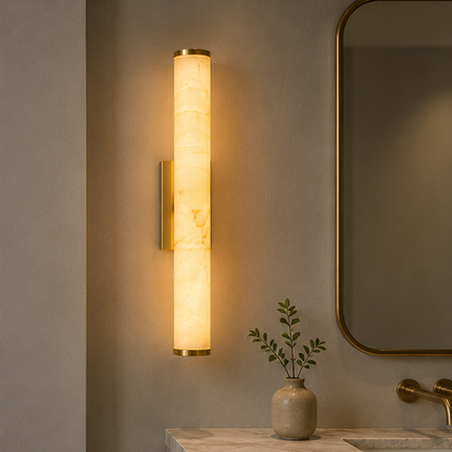 Leuchtende Wandlampe mit goldfarbenen Metallkappen über einem Waschtisch mit Marmorplatte, neben einem goldgerahmten Spiegel und einer Vase mit Zweigen. 2