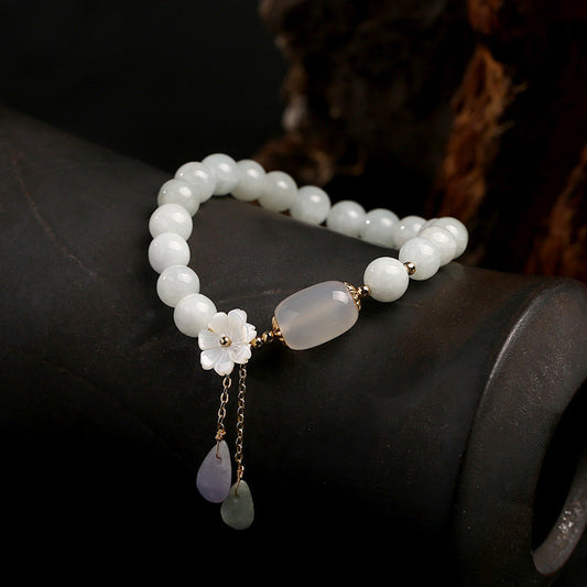 Jade-Armband mit weißen Perlen, Blumenanhänger und goldenen Akzenten auf schwarzem Hintergrund.