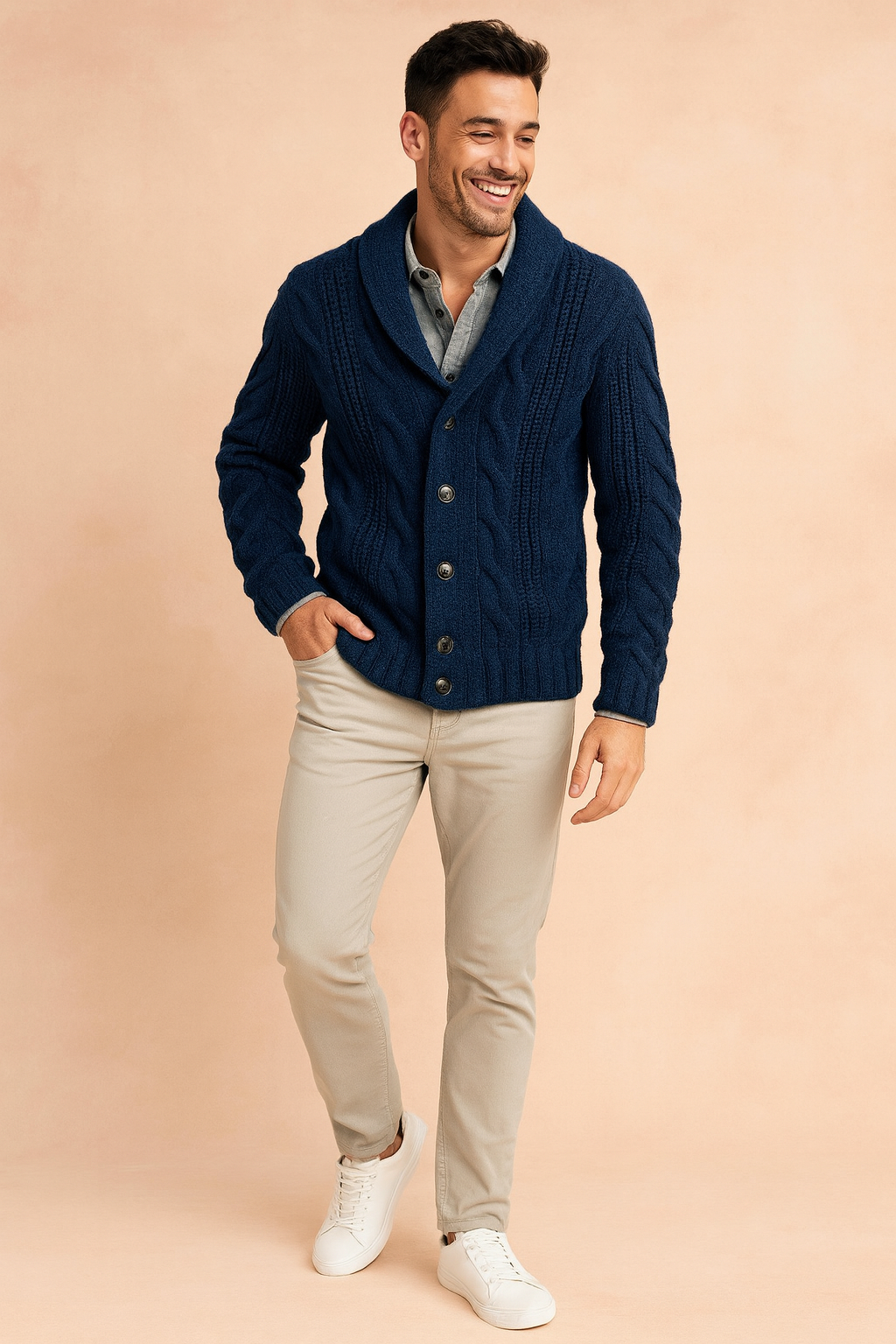 Ganzkörperaufnahme der marineblauen Strickjacke, kombiniert mit beiger Hose und weißen Sneakern vor pfirsichfarbenem Hintergrund. 2