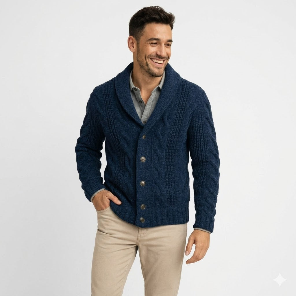 Lächelnder Mann in marineblauer Strickjacke mit Zopfmuster und Schalkragen, Hand in der Hosentasche, Halbfigur vor hellgrauem Hintergrund. 1