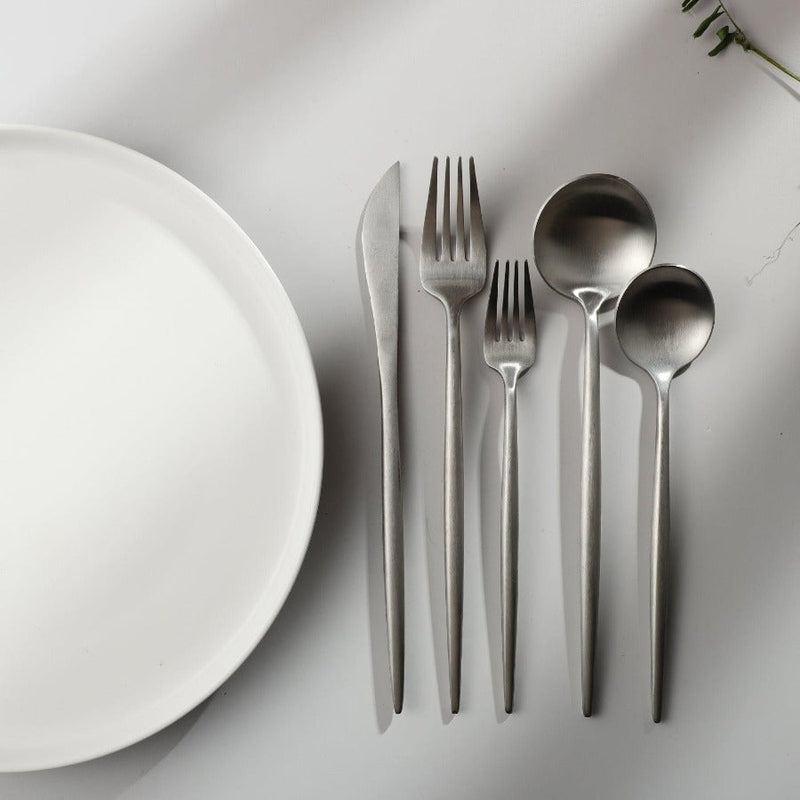 Minimalistisches Besteckset aus Edelstahl neben weißem Teller auf grauem Hintergrund.