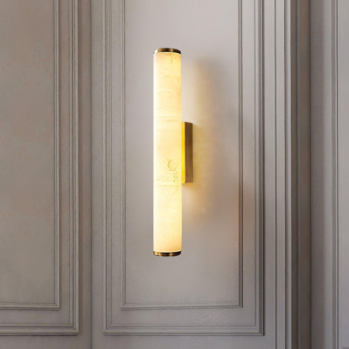 Wandlampe mit goldfarbenen Kappen und Halterung an einer hellen, mit Zierleisten verkleideten Wand, der Marmorzylinder leuchtet warm. 18
