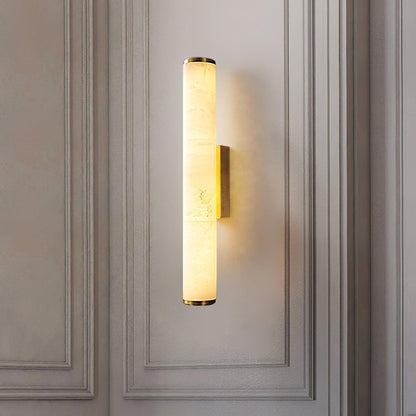 Wandlampe mit goldfarbenen Kappen und Halterung an einer hellen, mit Zierleisten verkleideten Wand, der Marmorzylinder leuchtet warm. 18