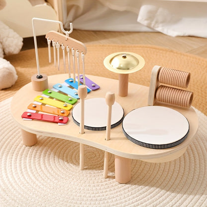 Musiktisch aus hellem Holz mit Regenbogen-Xylophon, Klangspiel, goldenem Becken, zwei Trommeln und Schlägeln auf einem Flechtteppich. 1