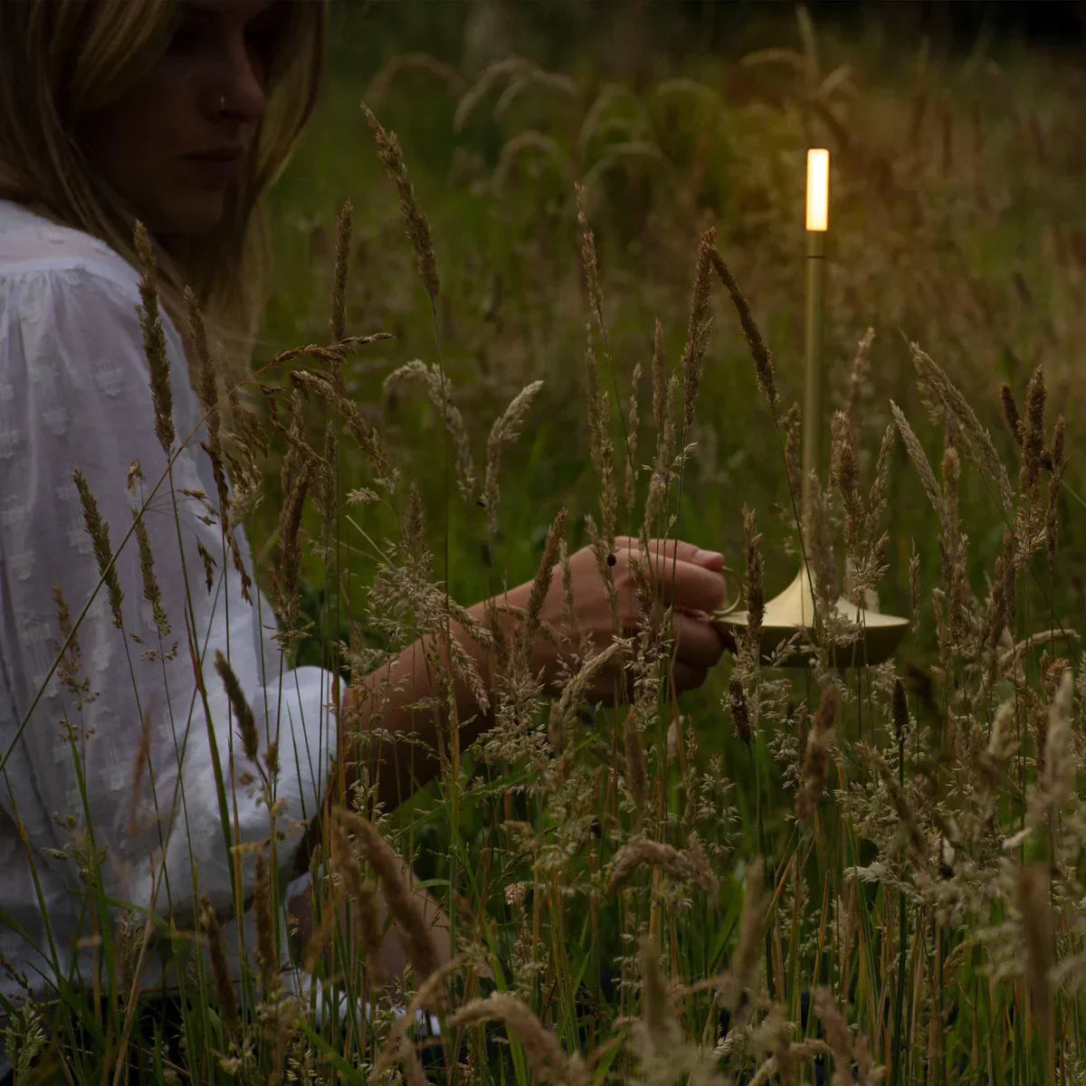 Frau in Wiese mit goldener, leuchtender Lampe, stimmungsvolle Beleuchtung, Natur, Abenddämmerung.