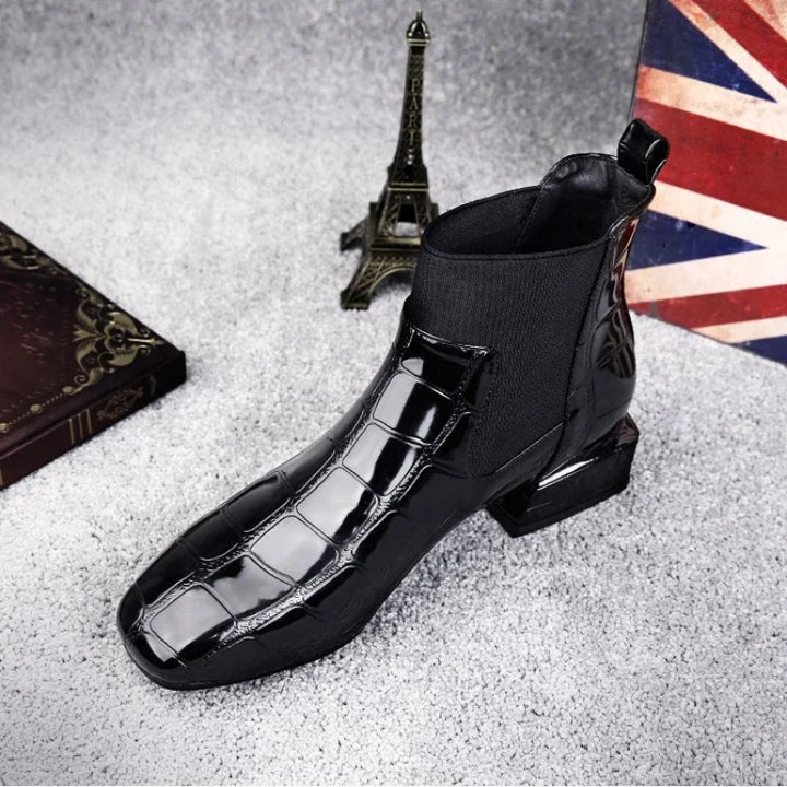 Schwarzer glänzender Stiefel mit Krokoprägung und Stretcheinsatz, leicht von oben vorn fotografiert, vor Eiffelturm-Figur und Union-Jack-Kissen. 1