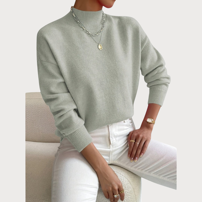 Pullover in zartem Graugrün mit Stehkragen, am Model zu weißer Jeans und Kettenschmuck, Sitzpose auf cremefarbenem Bouclé-Sessel. 2