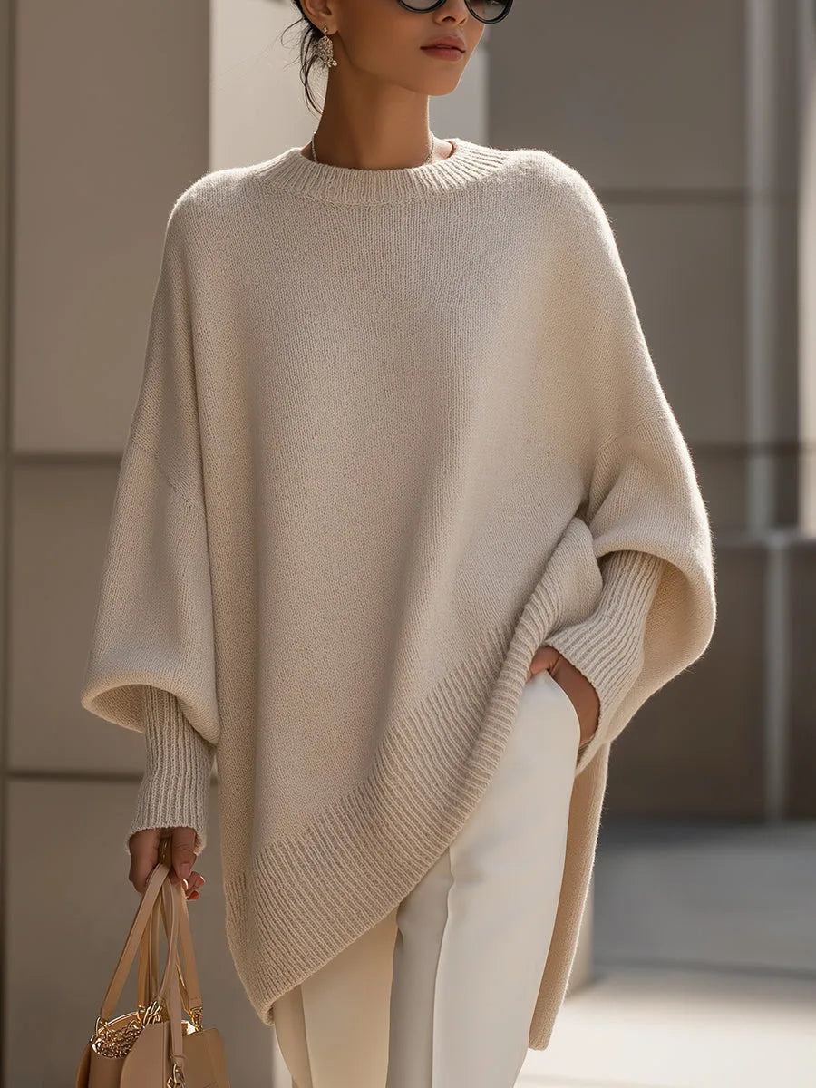 Beigefarbener Strickpullover mit weiten Fledermausärmeln, getragen von einer Frau mit weißer Hose, Handtasche und Sonnenbrille, Vorderansicht. 1