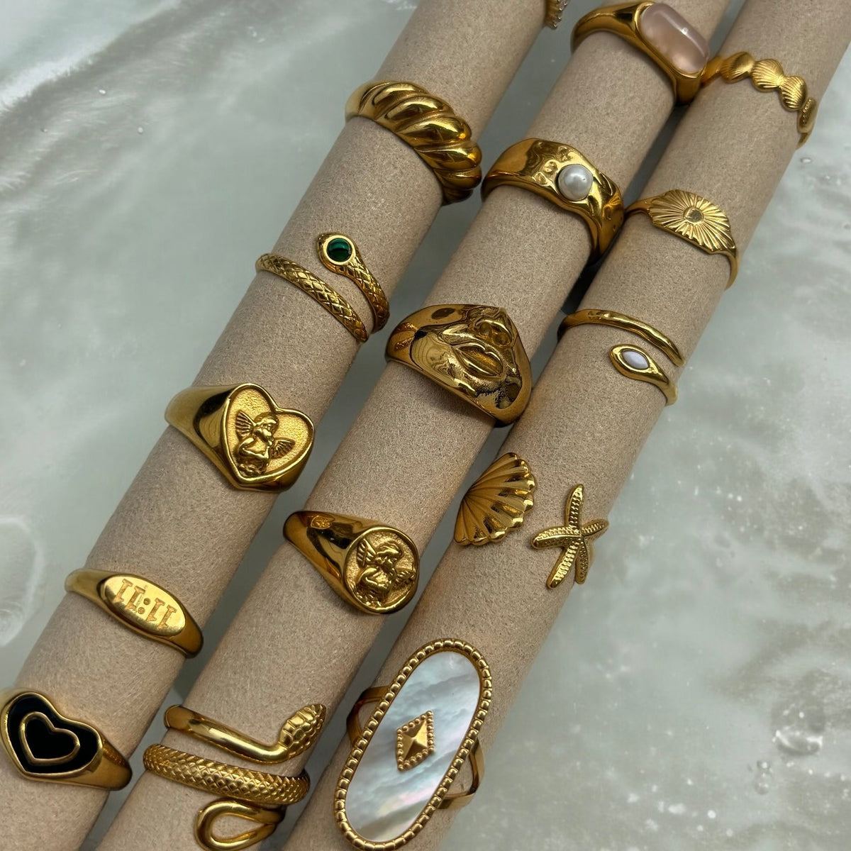 Flatlay mit dem Engelherz-Ring und vielen weiteren goldfarbenen Ringen, aufgesteckt auf beigen Rollen, darunter Schlangen- und Muschelringe. 5