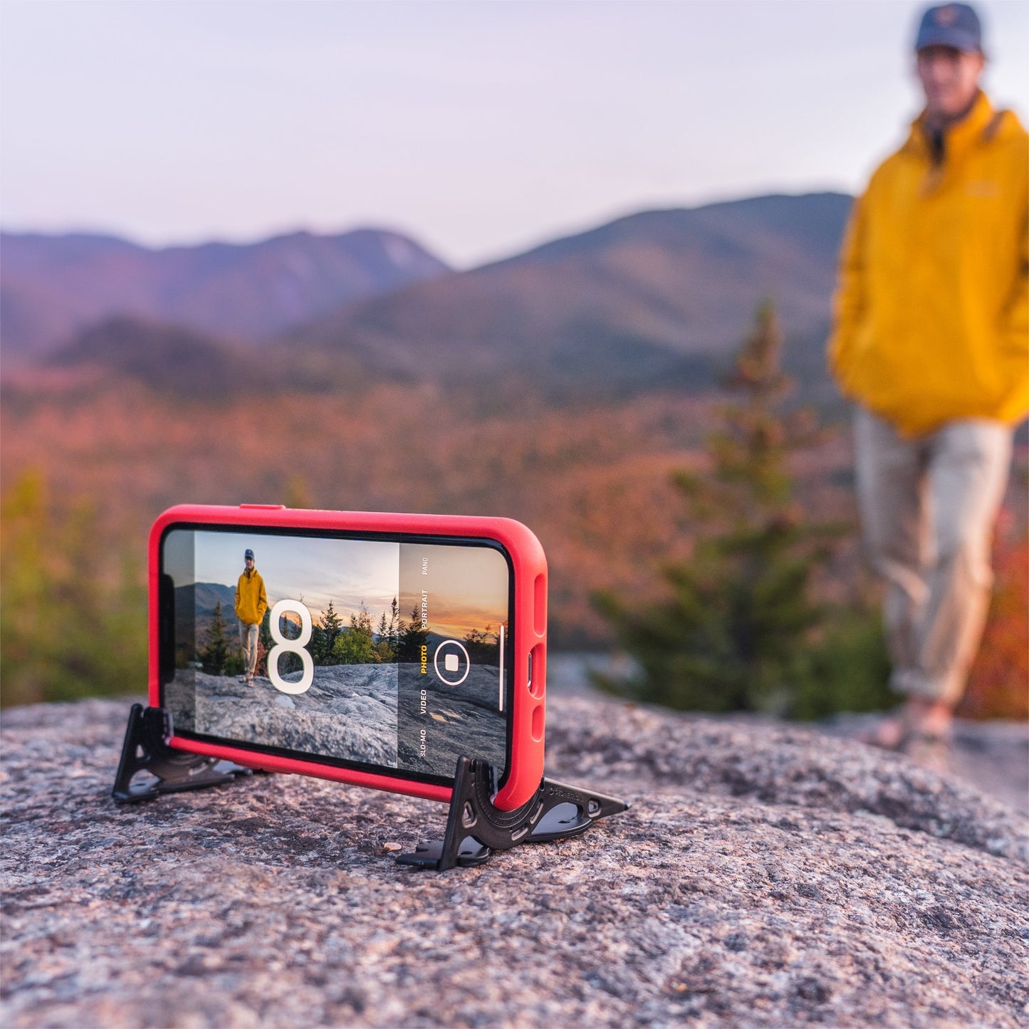 Rotes Smartphone im PocketTripod auf Felsen, Herbstlandschaft, Mann im Hintergrund.