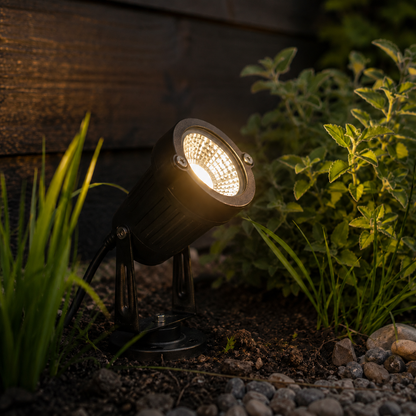 Leuchtender Gartenstrahler in der Dämmerung, schräg im Erdboden zwischen Gras, Kräutern und Kieselsteinen vor einer Holzwand, mit sichtbarem Kabel und Bodenplat 6