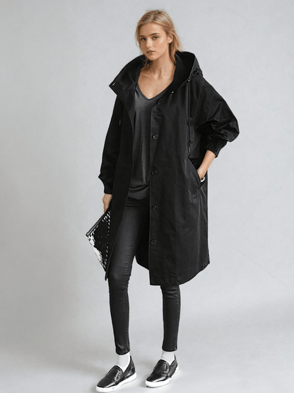 Ganzkörperansicht in Schwarz: Der Mantel mit Kapuze und Knopfleiste, kombiniert mit schwarzem Top, enger Hose, Slippern und Clutch. 2