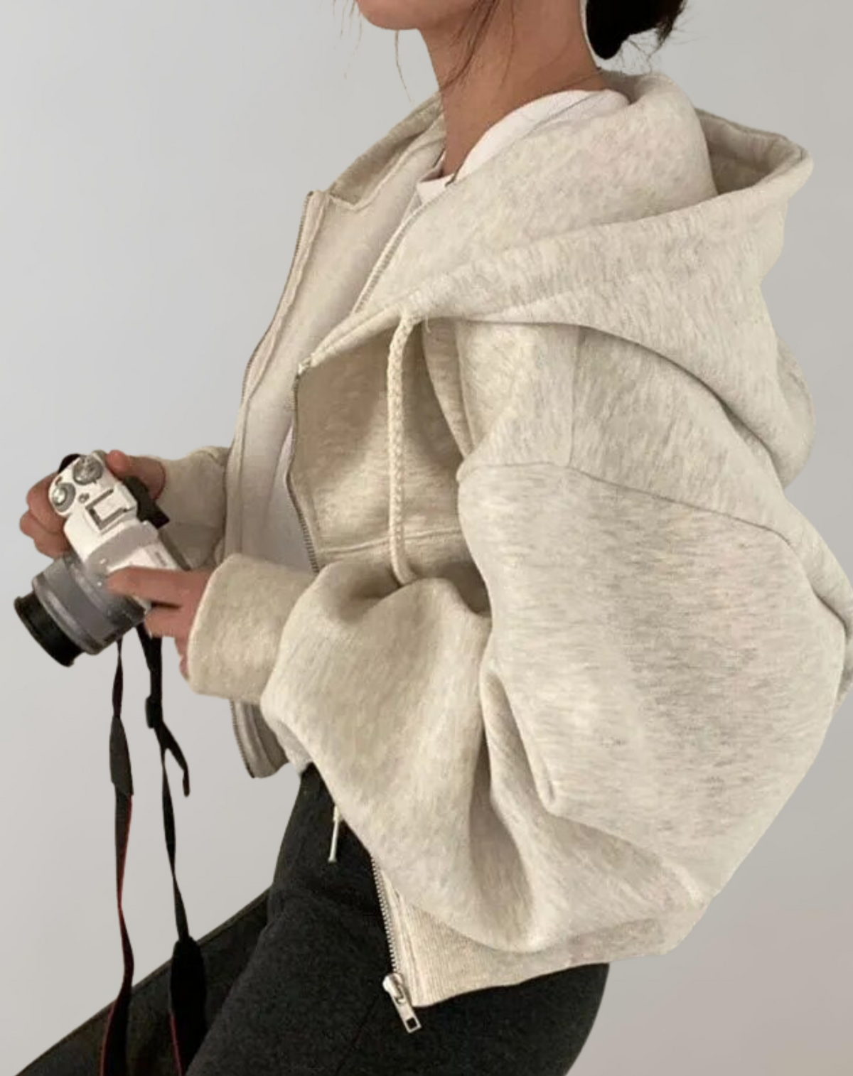 Helles, meliertes Hoodie-Modell in Seitenansicht mit weit geschnittenem Ärmel und Kapuze, die Trägerin hält eine Kamera in den Händen. 2