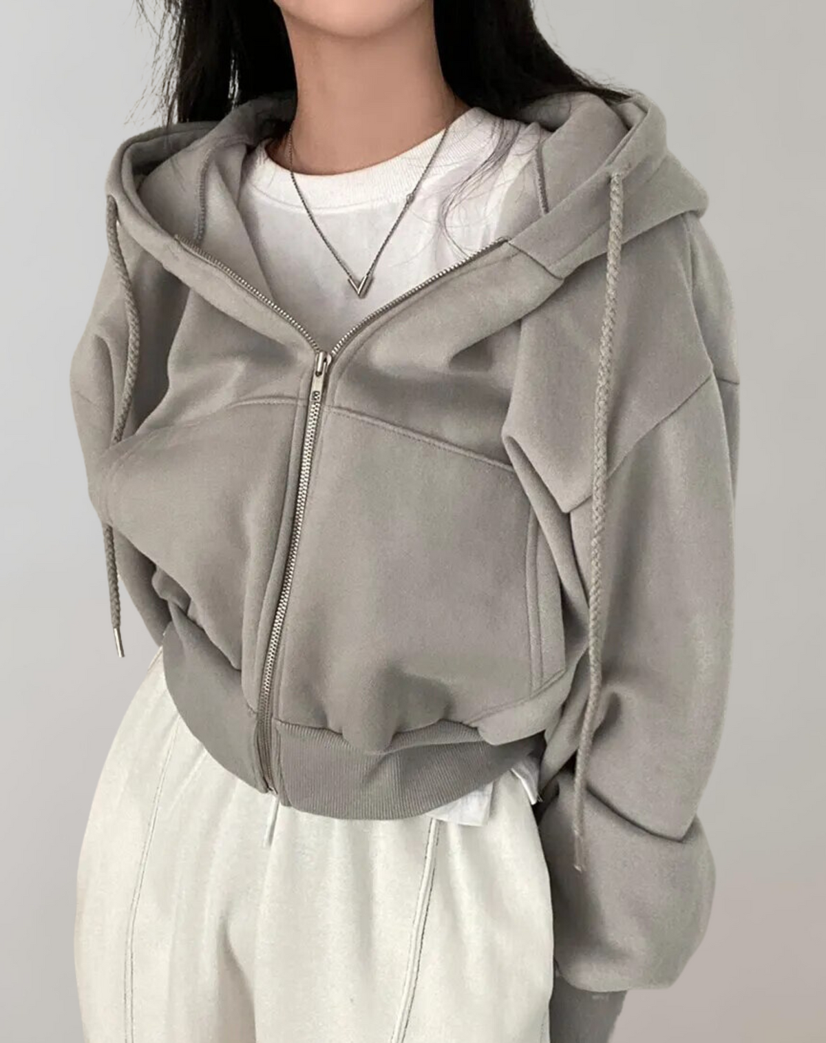 Grauer Hoodie mit Reißverschluss, Kordeln und Rippbündchen am Saum, kombiniert mit weißem Shirt, Halskette und heller Hose. 4