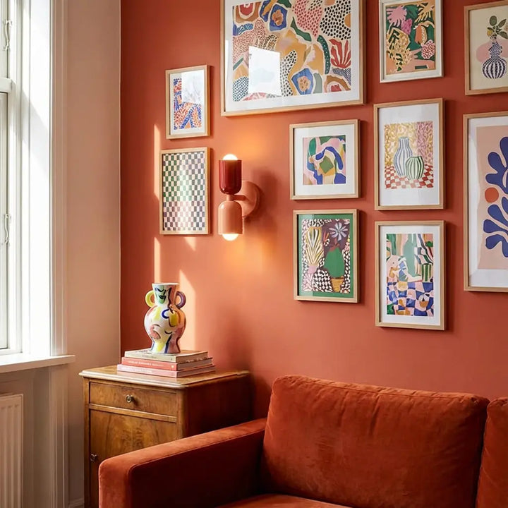 Wandleuchte mit dunkelrotem und rosa Schirm an einer Terrakottawand zwischen bunten Bildern, darunter ein Samtsofa und eine Kommode. 5