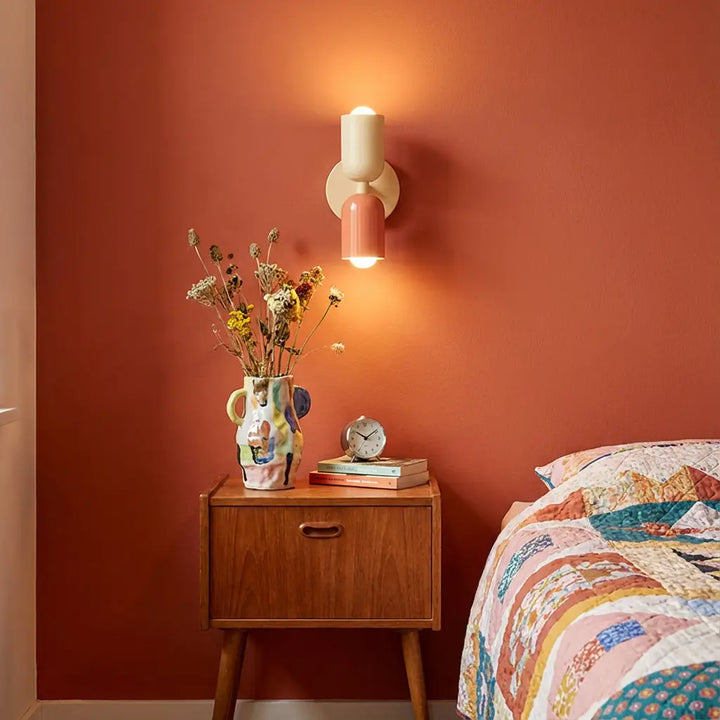 Leuchtende Wandleuchte in Creme und Lachsrosa an terrakottafarbener Wand über einem Holz-Nachttisch mit Vase, Büchern und Wecker. 1