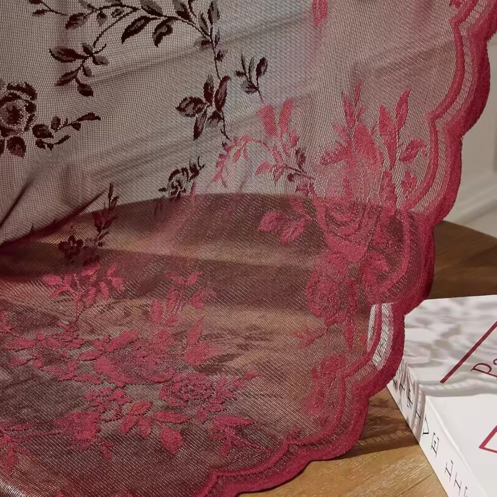 Nahaufnahme des weinroten Stoffs mit gewelltem Saum und aufgewebten Rosenranken, der locker über einer Holzfläche neben einem Buch liegt. 3