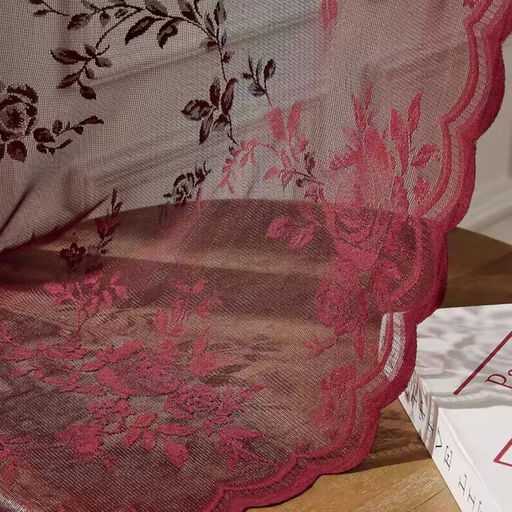 Nahaufnahme des weinroten Stoffs mit gewelltem Saum und aufgewebten Rosenranken, der locker über einer Holzfläche neben einem Buch liegt. 3