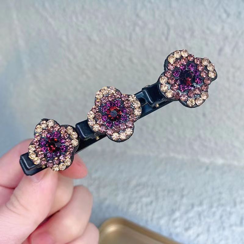 Detailansicht einer schwarzen Haarspange mit drei Strassblüten in Violett-, Rosé- und Goldtönen, vor heller Wand gehalten. 2
