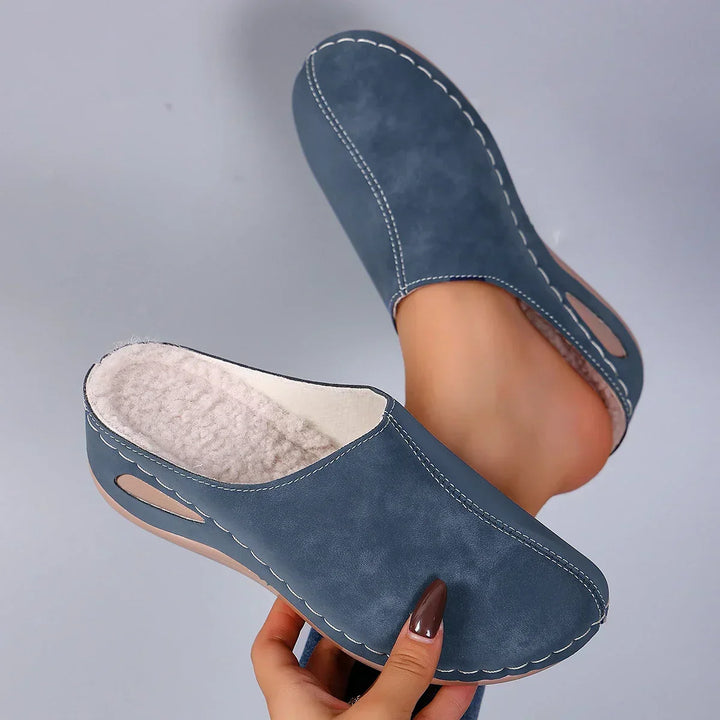 Blaue geschlossene Pantoffeln mit weißen Ziernähten, Fleecefutter und seitlicher Aussparung, von einer Hand hochgehalten, ein Fuß steckt darin. 1
