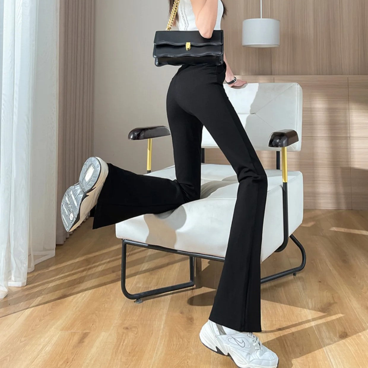Frau in schwarzer Schlaghose und Sneakers, hält schwarze Handtasche, modernes Interieur.