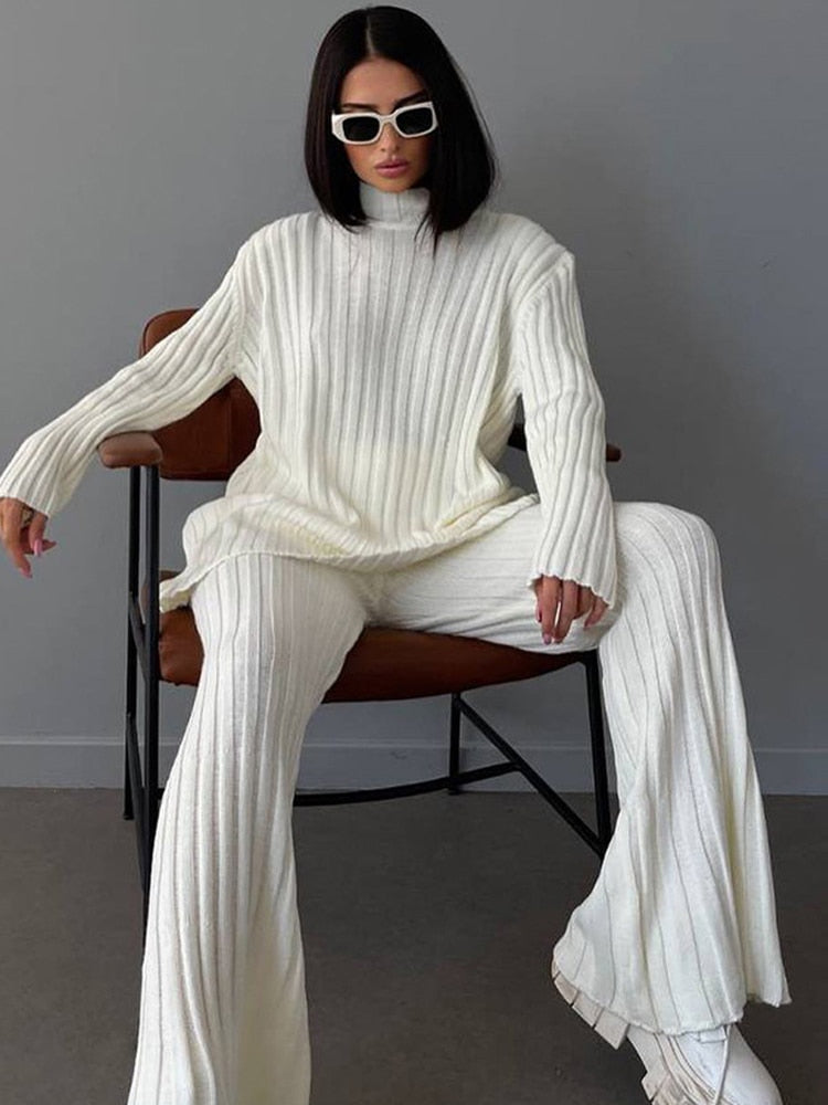 Frau in weißem Strickpullover und Hose, sitzt auf Stuhl, trägt Sonnenbrille.