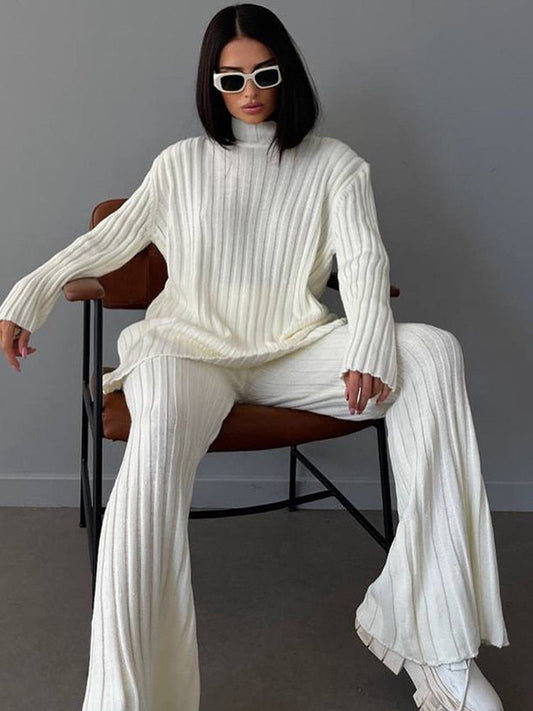 Frau in weißem Strickpullover und Hose, sitzt auf Stuhl, trägt Sonnenbrille.