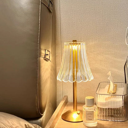 Goldene Tischlampe mit Glaslampenschirm auf Nachttisch, neben Parfümflasche und Taschentuchbox.