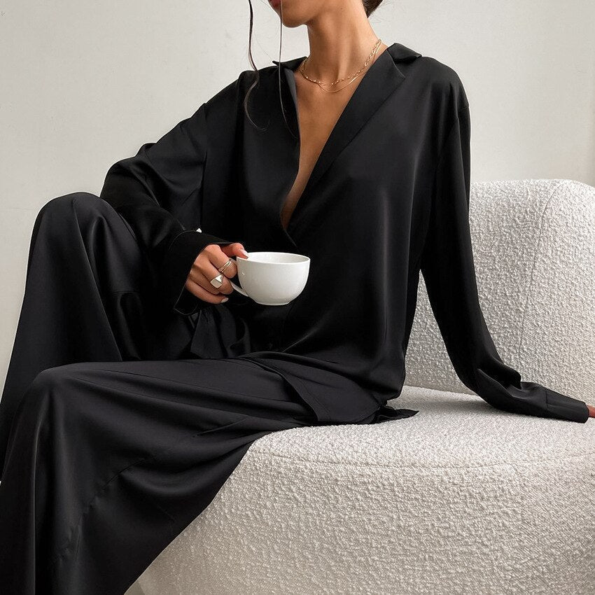 Frau in schwarzem Satin-Pyjama hält weiße Tasse, sitzt auf modernem Sessel.