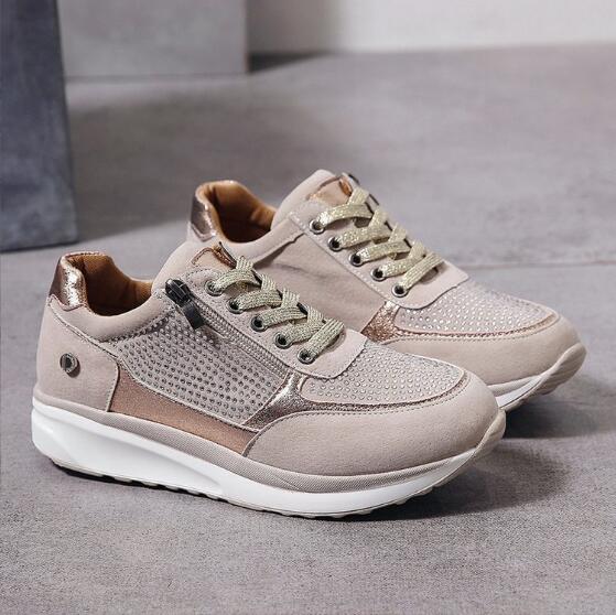 Paar beigefarbene Sneaker mit goldfarbenen Glitzerschnürsenkeln, Strassbesatz und weißer Sohle, auf grauem Boden fotografiert. 2