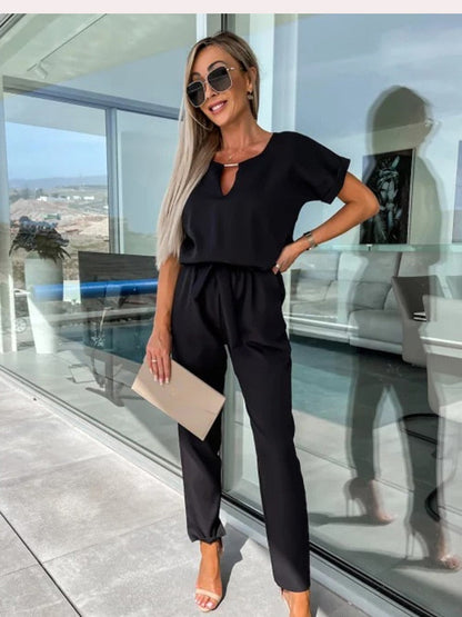 Frau in schwarzem Jumpsuit, Sonnenbrille, hält beige Clutch, moderne Terrasse im Hintergrund.