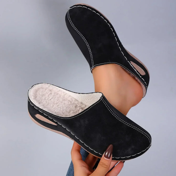 Schwarze Pantoffeln von oben gezeigt, das helle Fleecefutter ist im Inneren zu sehen, ein Fuß steckt im zweiten Schuh. 5