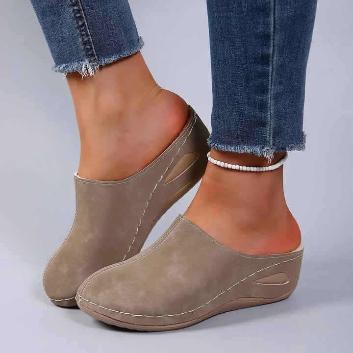 Beigefarbene Pantoffeln mit Keilsohle an den Füßen einer Frau mit Jeans und weißer Perlenkette am Knöchel. 3