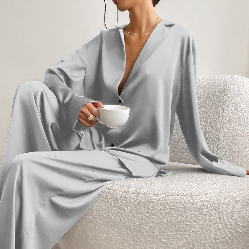 Frau in grauem Satin-Pyjama hält Tasse, sitzt auf weißem Sofa, elegantes Lounge-Outfit.