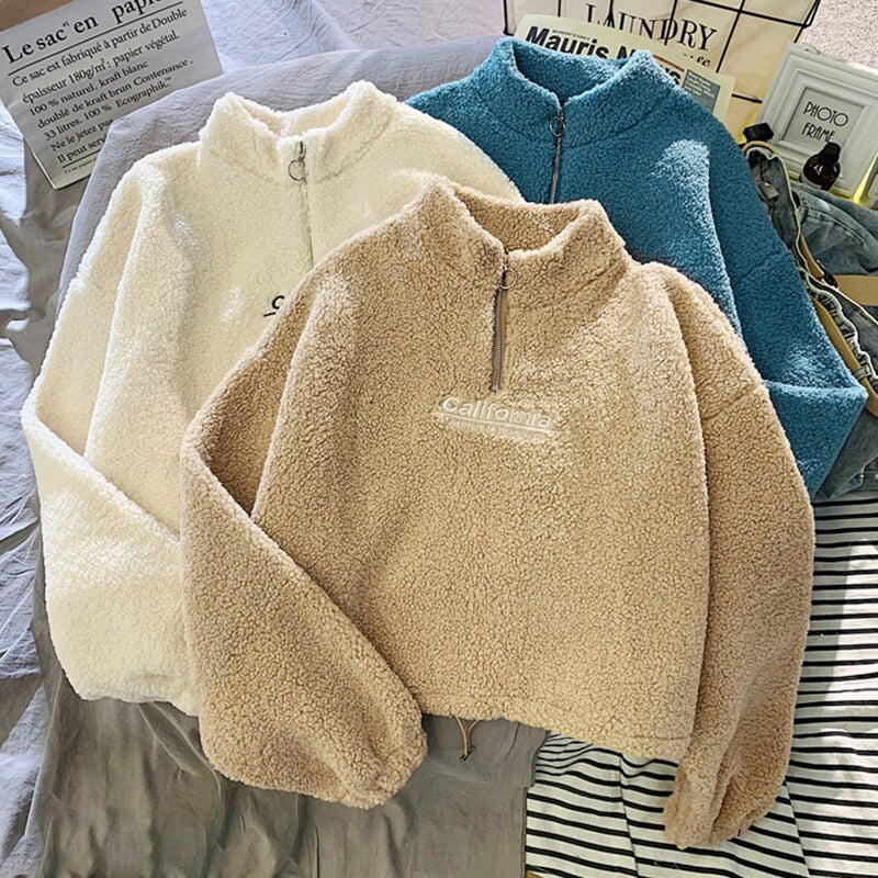 Drei flauschige Fleece-Pullover in Weiß, Beige und Blau mit Reißverschluss auf gestreiftem Hintergrund.