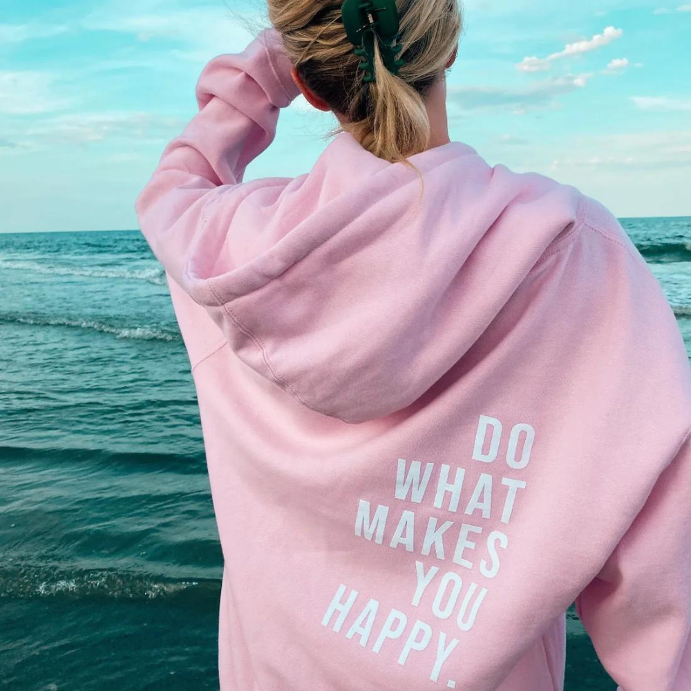Frau in rosa Hoodie mit Aufschrift am Strand, Meer im Hintergrund, Freizeitmode, Sommer.