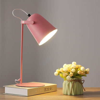 Rosafarbene Tischlampe mit leuchtendem Schirm auf einem Buch, daneben eine Vase mit hellen Röschen auf einem Holztisch. 8