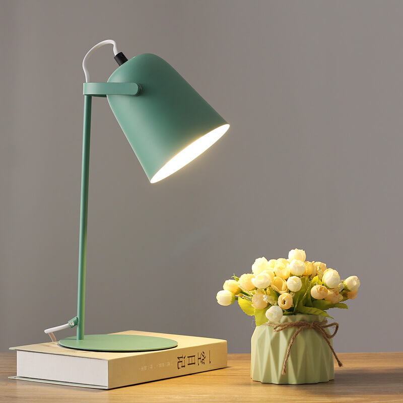 Grüne Tischlampe mit eimerförmigem Schirm, eingeschaltet auf einem Buch, daneben eine kleine Vase mit hellen Röschen. 9